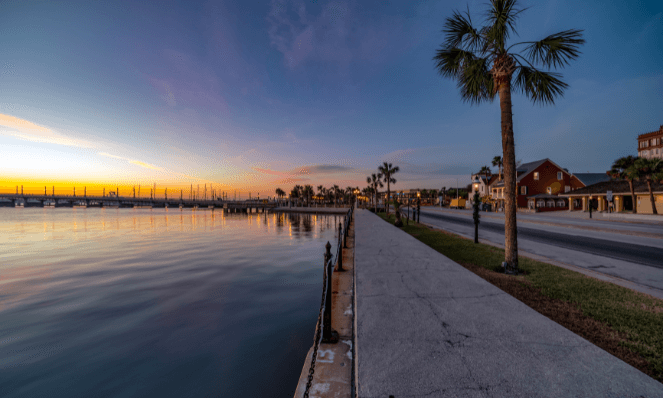 View of Historic St. Augustine downtown during sunset hours with yellow and blue skies, palm trees, water, and buildings lining the side of the street