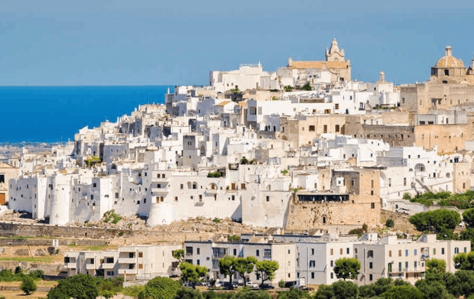 an aerial view of Ostuni, Italy with white washed buildings, trees, and a blue sea