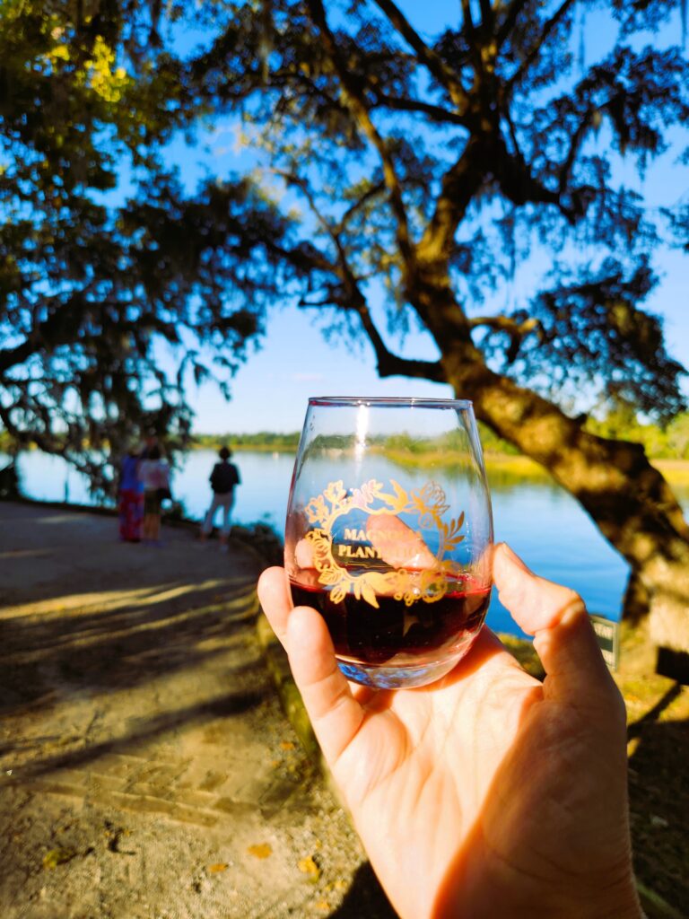 A hand holding up a stemless signature glass at one of the Magnolia Plantation and Gardens private evening Wine Strolls with the view of the Ashley River at sunset in the background.