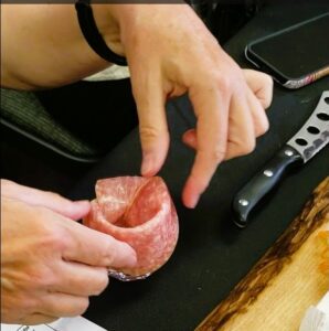 Hands creating a meat rose around a small glass, the charcuterie board and a cheese knife in background