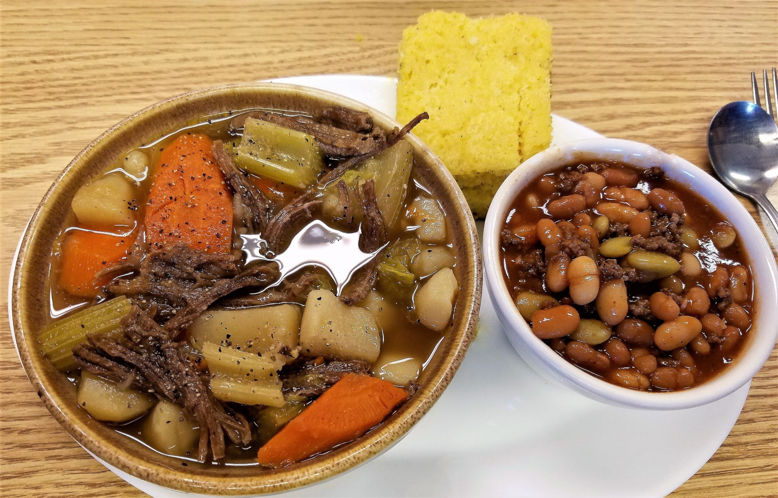 A bowl of meat, carrots, and potatoes with cornbread, and a small bowl of beans with meat mixed throughout.