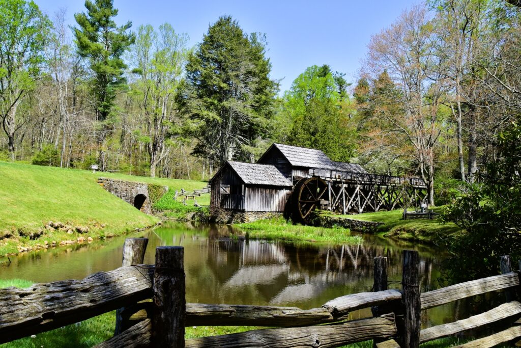 A weathered gray water-powered grist mill on the Blue Ridge Parkway, surrounded by a weathered split-rail fence, and the mill reflects in the pond.