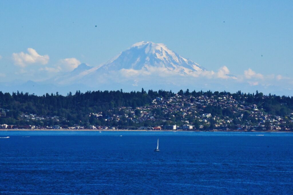 Snow-topped Mount Ranier rising in the background, with the town of Seattle on the hillside, surrounded by water and a sail boat in the foreground.