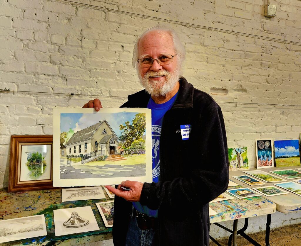 White-haired, bearded gentleman holding a painting of a LaGrange church he created with many other paintings in the background