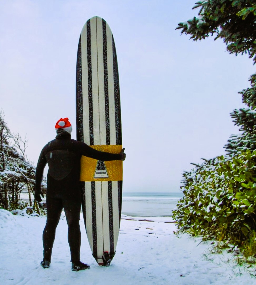 Man in wet suit and Santa hat ready to hit the waves to surf, holding his surf board looking out at the snow-covered beach