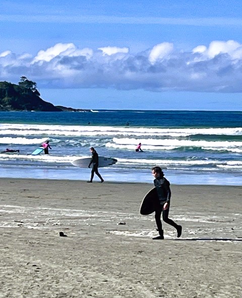 Surfers in the dark blue ocean, with two surfers carrying their boards across Tofino's Cox Bay sandy beach