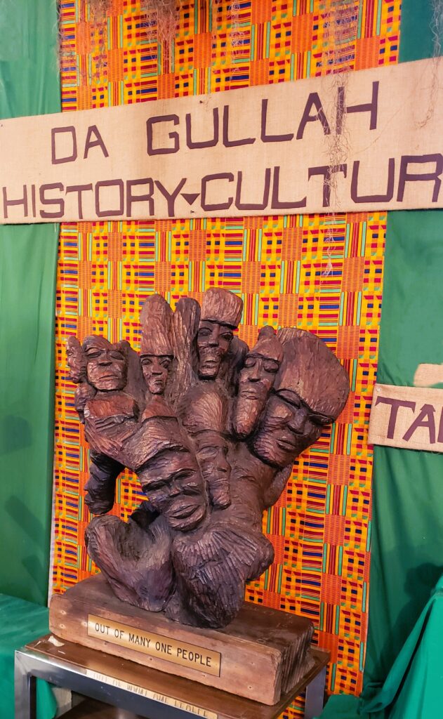 African sculpture with the title: "Out of many, one people" in front of bright African print fabric of green, yellow, purple, and orange. Gullah history is kept alive in historic Georgetown SC.