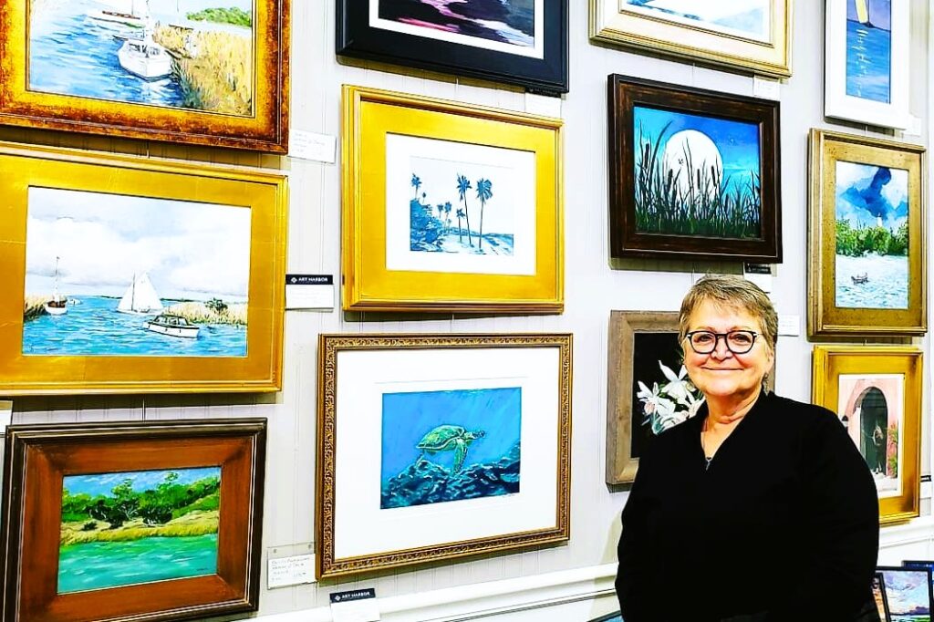 Artist and Gallery owner Deb, standing in front of a wall of framed paintings, ready for sale.