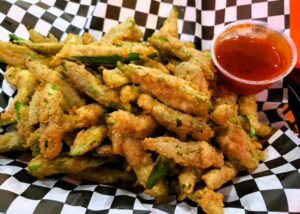 Battered and fried strips of okra, on Black & white checked serving paper and a container of Roasted Pepper Bacon Jam