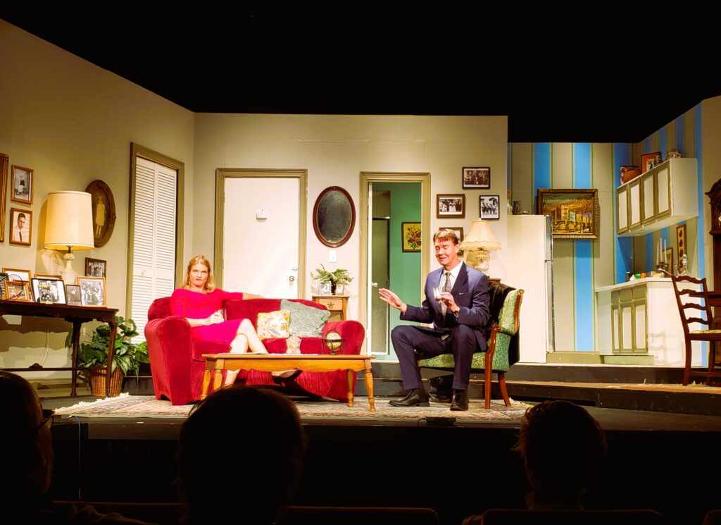 A female cast member sitting on a love seat, and a male actor sits in a chair in a living room set on stage at The Strand Theatre.