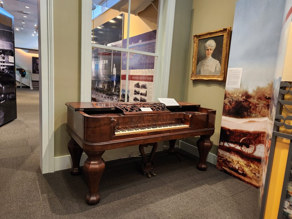 A museum display showing photos,then an oil portrait of a woman, and next, a mahagony spinet piano