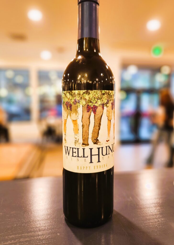 A bottle of wine, showing the Well Hung Vineyards label, men wearing pants, and covered with grapevines.