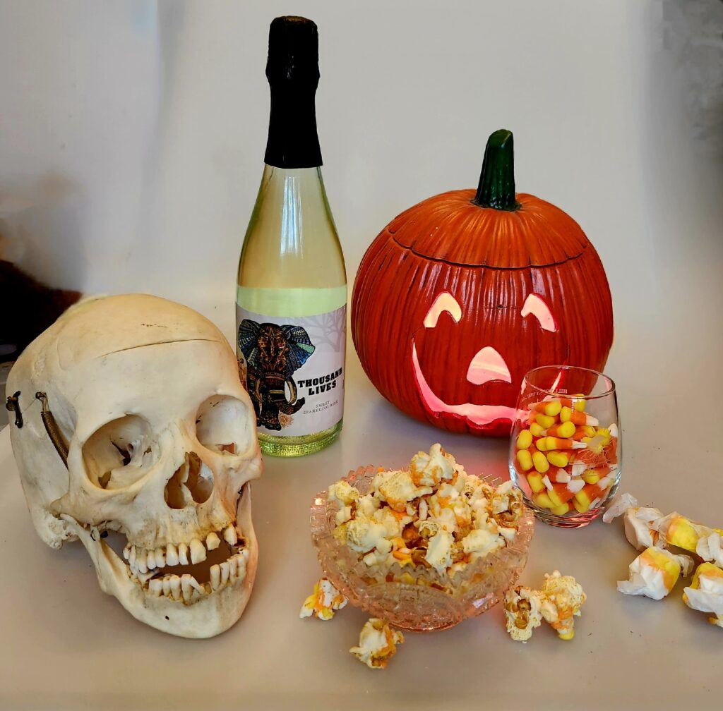 Skull and lighted jack-o-lantern with bottle of white bubbly, candy corn, popcorn drizzled with melted candy corn, and candy corn taffy.