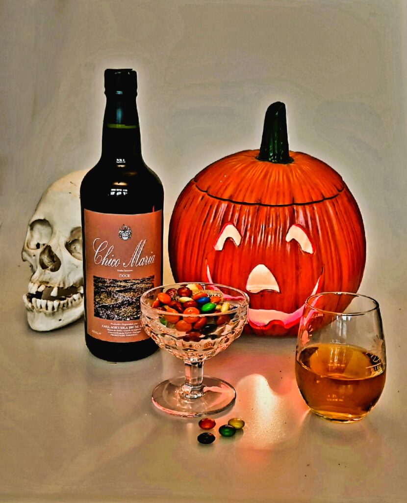 A bottle of Port and an antique glass goblet filled with M & Ms, alongside a glass of port in a display with a ceramic jack-o-lantern and skull.