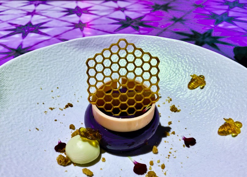 Lavender Honey dessert on white china plate, which is topped with edible honey comb, and then surrounded by edible honey bees and edible flower petals.