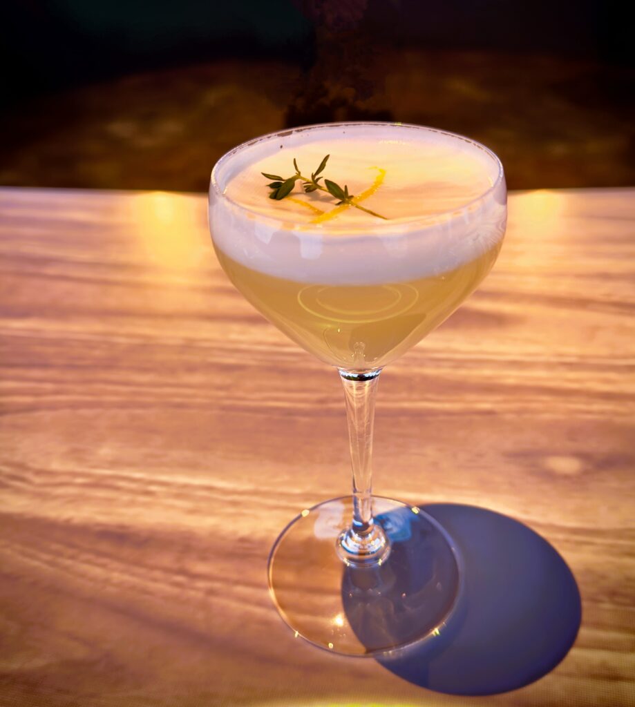 A pale yellow drink topped with whipped egg whites, and a piece of lemon peel and rosemary floating on top.