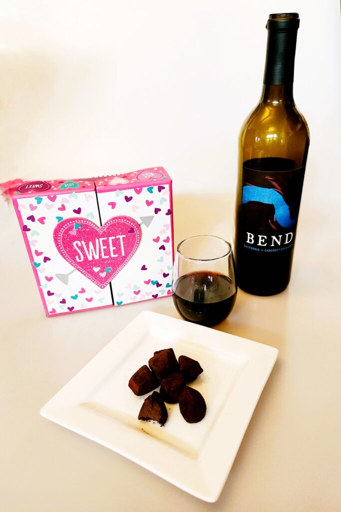 Wine and Chocolate photo shows bottle of Bend Cab Sav, a stemless glass of red wine, a plate of dark chocolate truffles, and a Valentine's Day Gift Box