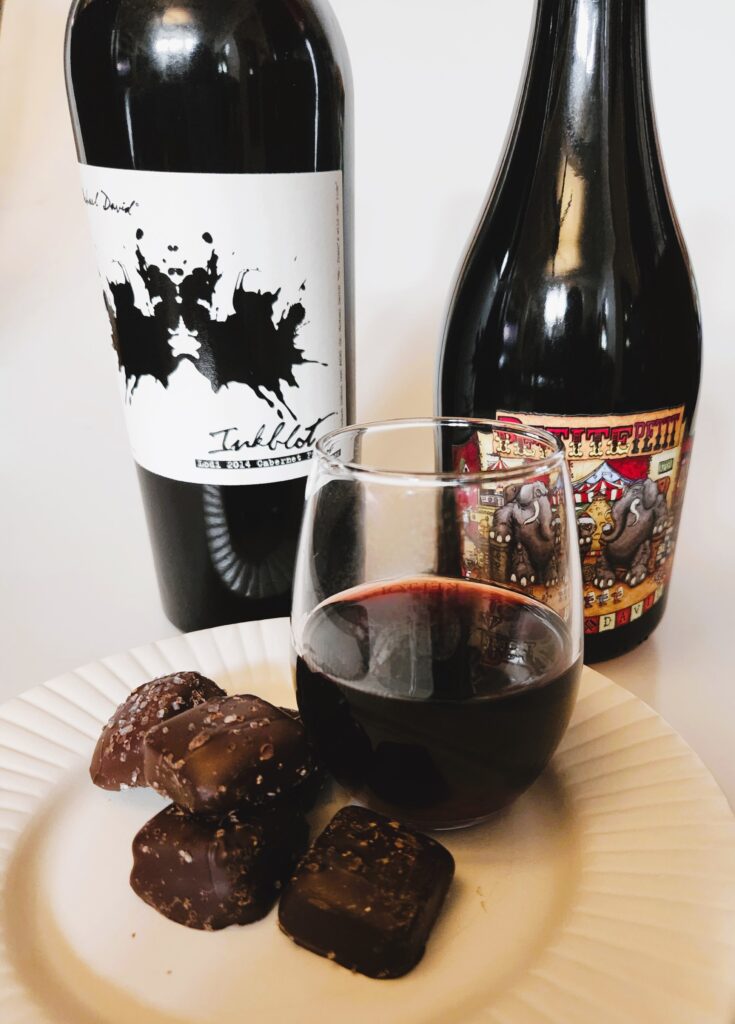 Wine and Chocolate photo shows bottles of Petit Syrah and Cab Sav, red wine in a stemless glass, and dark chocolate sea salt caramels.