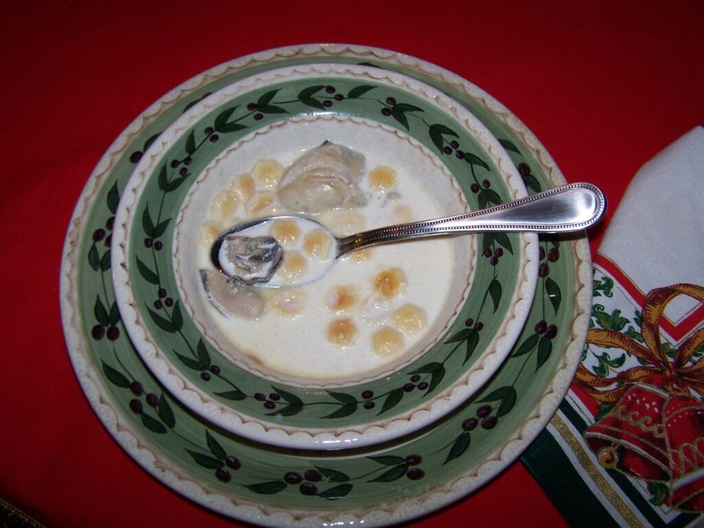 A plate and bowl with green trim and olive leaves with olives, the bowl filled with oyster stew and oyster crackers. Sitting on a red tablecloth with a Christmas napkin to the side.