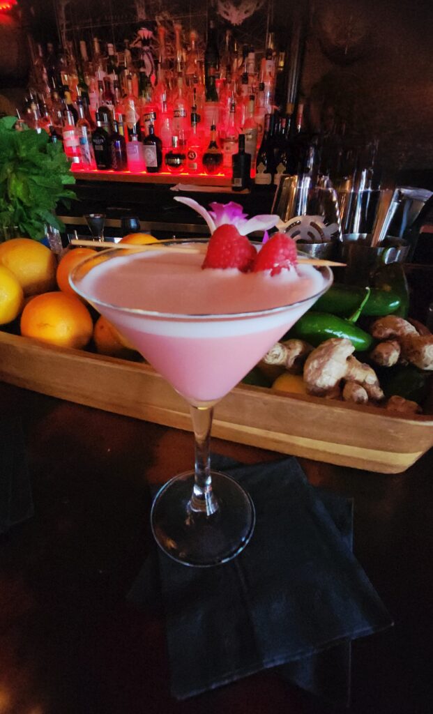 A pink drink topped with foam (from egg whites) and two raspberries on  a skewer with a purple orchid bloom.