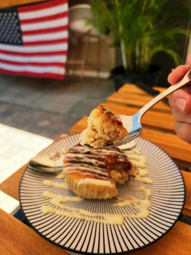 A bite of Crème Brûléed Bread Pudding on a fork, held over the entire serving on a blue and white striped plate, drizzled with frosting.