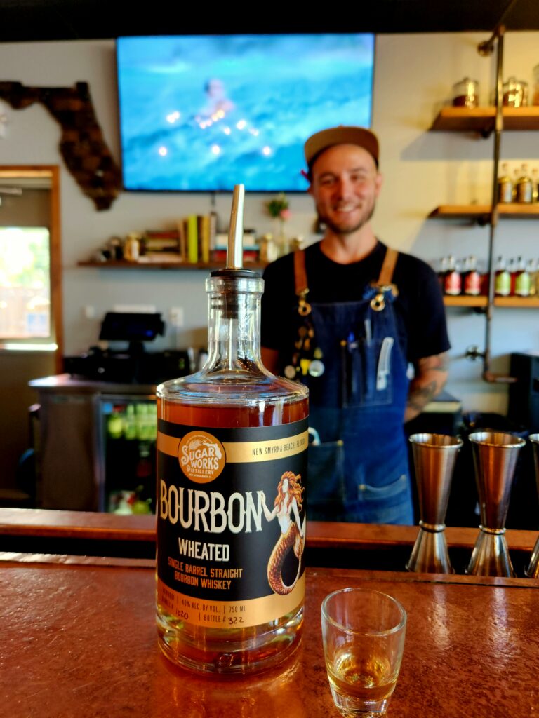 Bartender standing behind bar at Sugar Works in New Smyrna Beach, with a bottle of wheated bourbon on the bar.