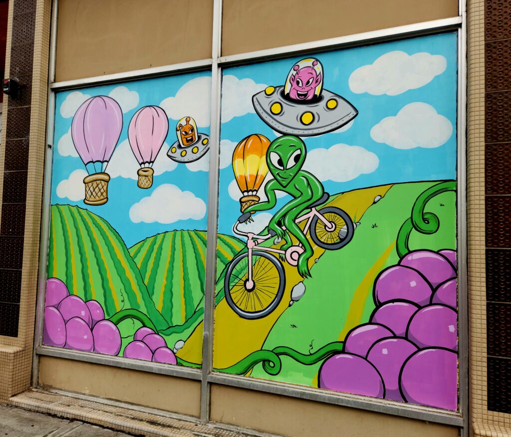 An alien from outer space on a bike crossing the hills of a vineyard, while spaceships and hot air balloons hover above.