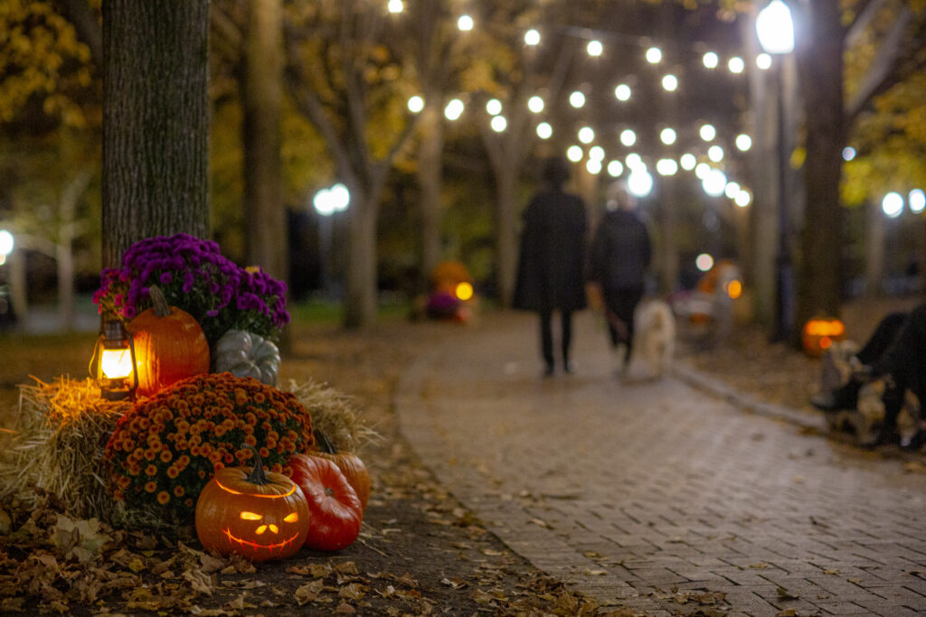A couple walk along a brick path lined with lanterns, incandescent lights, and carved jack-o-lanterns.