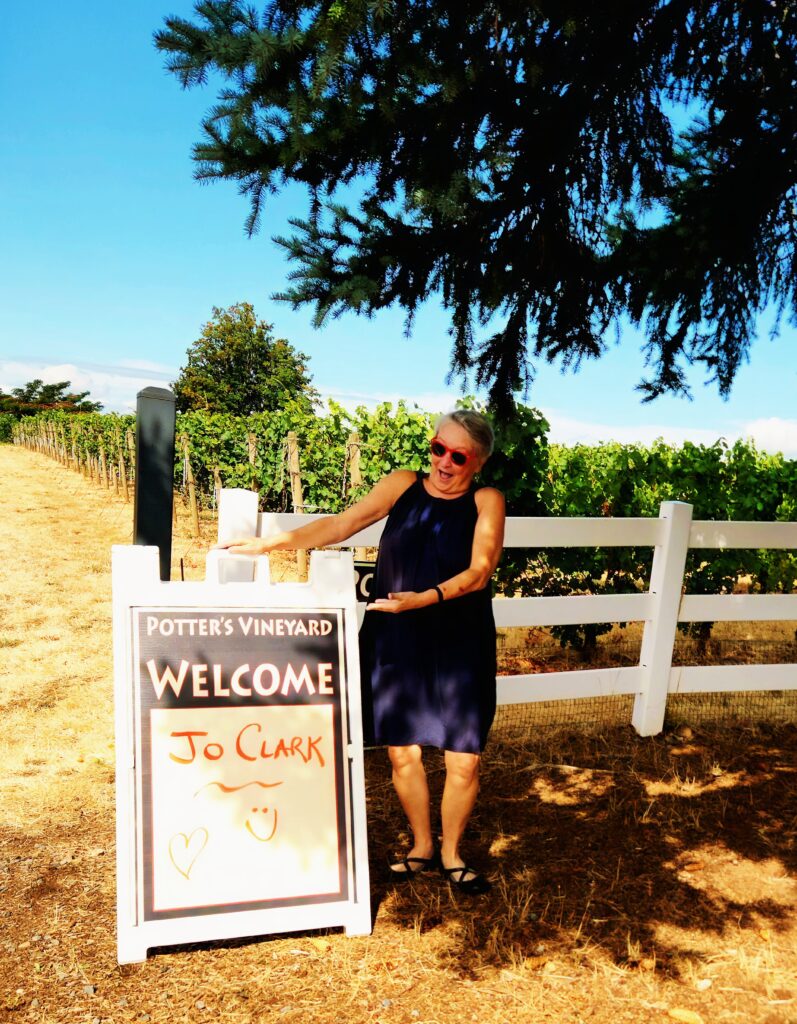 26 Sensational Must-Visit Oregon Wineries in the Willamette Valley 30 Woman in front of vineyard beside sign that says .WELCOME Jo Clark