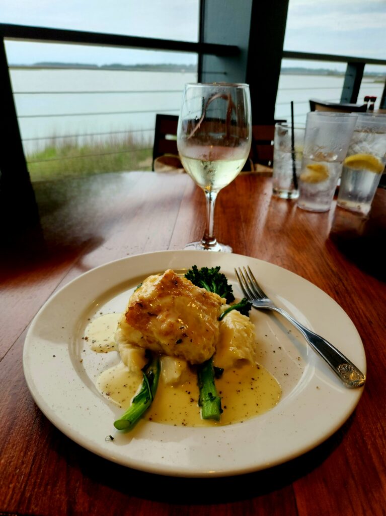 2 Islands, a Lighthouse, & Secret Magic of the Lowcountry 35 Glass of white wine with a piece of fish, atop asparagus spears and mashed potatoes, with a view of the water in the background.