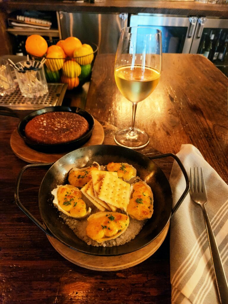 2 Islands, a Lighthouse, & Secret Magic of the Lowcountry 65 A plate of oysters on the half-shel, iron skillet of cornbread, and a glass of white wine, gray and white napkin beside the dish topped with a fork.
