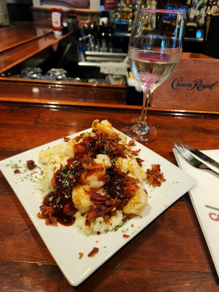 2 Islands, a Lighthouse, & Secret Magic of the Lowcountry 66 Shrimp and bacon topping grits and dripping with BBQ sauce on a white square plate.