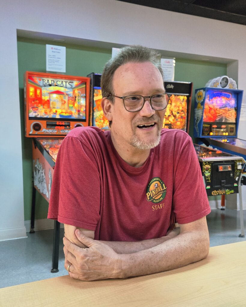 Greensboro: Big, Unexpected Adventures From 1781 Battlefield to Jeansboro 34 Man wearing Pinball Museum t-shirt with pinball machines in background.
